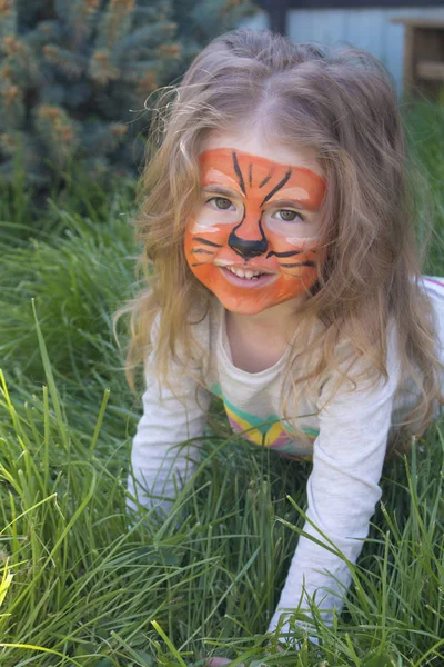 Porträtt av en liten flicka med tiger aqua makeup, leende och sitter i en park. — Stockfoto