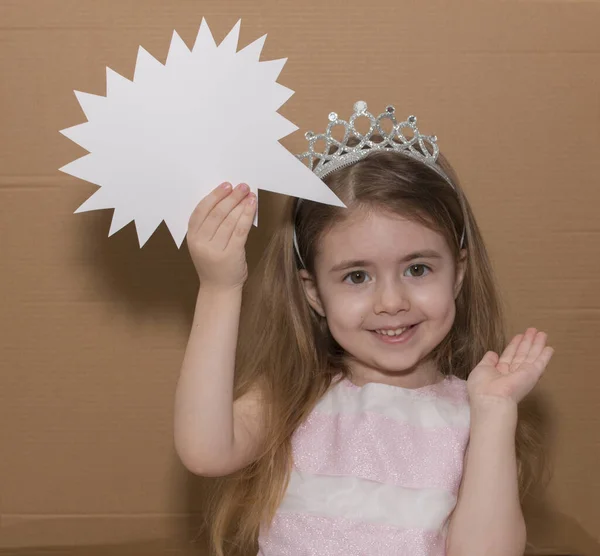 Imagen de niña confundida con una corona de pie aislada sobre fondo de cartón gris sosteniendo la burbuja del habla. Mirando cámara. espacio para texto — Foto de Stock