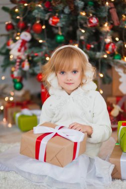 Onun ellerinde hediyelerle yılbaşı ağacının yanında oturan küçük güzel kız. Beyaz bir kazak ve sıcak kulaklıklar