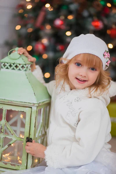 Küçük güzel kız beyaz bir şapka ve kıyafet Noel ağacının yanında oturur