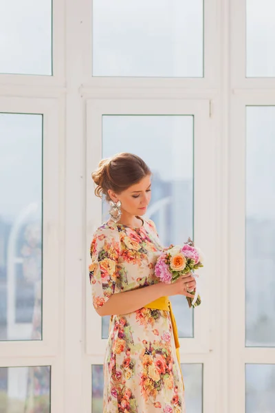 Bella ragazza in un abito lungo in piedi vicino a una finestra con un bouquet — Foto Stock