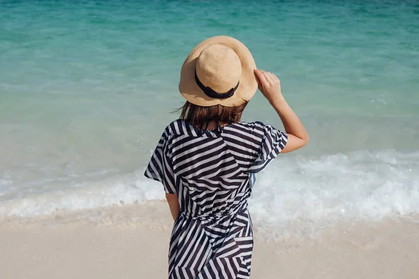 在大海的背景上，一个戴着帽子，穿着条纹连衣裙的少女在海滩上。泰国，竹岛 免版税图库图片