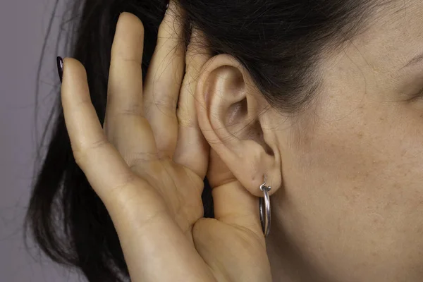 A closeup and side profile view of a young caucasian woman struggling to hear the conversation, gesturing with her hand behind her ear to speak up, wax build up and deafness.