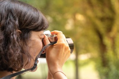 Beyaz bir kadının doğal ortamda fotoğraf çekerken çekilmiş yakın bir görüntüsü. Ormandaki bir manzara fotoğrafını çerçevelerken yüzüne bir kamera tutuyordu. Boşluğu sağda kopyala.