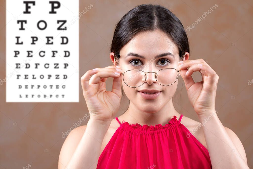 A stunning young Caucasian girl with brunette hair is seen trying on new spectacles at the optometrist. Front portrait view of lady with soft facial features at visit to eye doctor.