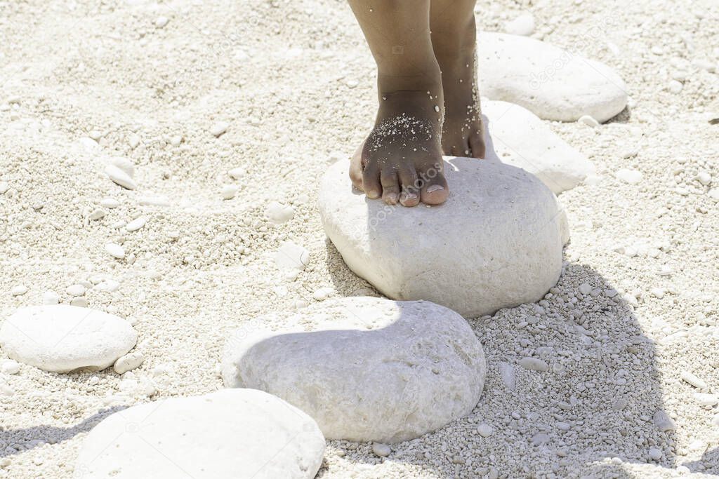 A closeup view on the bare feet of a young child playing and standing on white stones on a sandy beach during summer vacation, stepping stones with room for copy.
