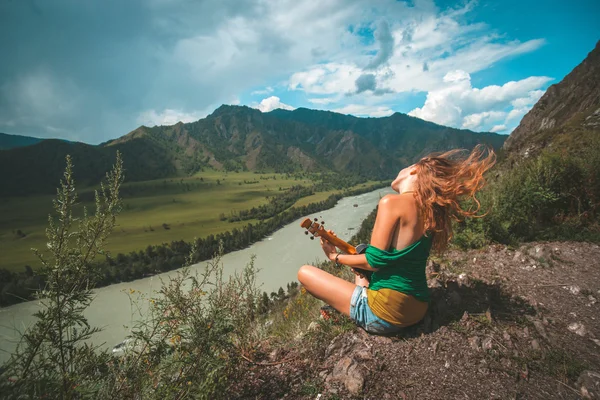 Fiatal vörös hajú nő játék ukulele a hegy csúcs. Hegyi folyó táj. — Stock Fotó