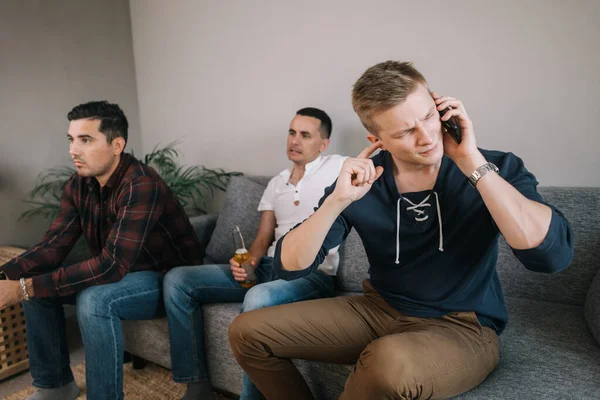Guy trying to talk on the phone. Behind the man back his friends are watching TV