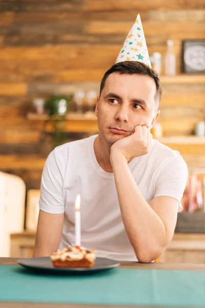 Upset young man sitting at the birthday cake and looking with sad eyes on it.