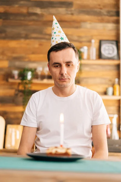 Sad lonely young man celebrating birthday alone, sitting at the birthday cake.