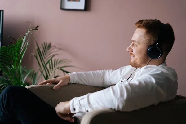 Young relaxed man in headphones sitting on soft chair in living room