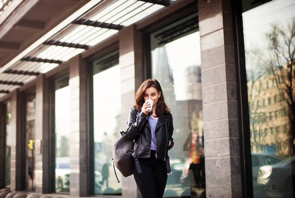 Ung vacker brunett dricka kaffe promenader runt city.leather jacka, urban ryggsäck, röda läppar — Stockfoto