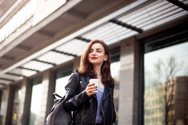 Ung vacker brunett dricka kaffe promenader runt city.leather jacka, urban ryggsäck, röda läppar — Stockfoto