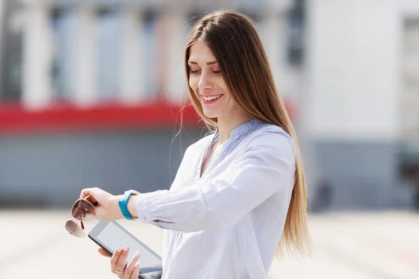 Wesoły młoda kobieta piękne w niebieska koszula utrzymuje jej touchpad z uśmiechem i patrzy na zegarek — Zdjęcie stockowe