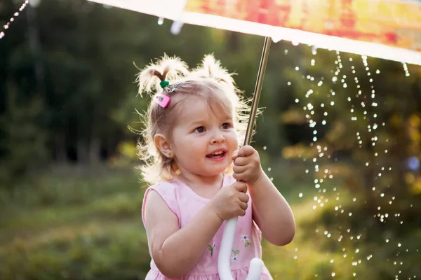 Pequeña chica alegre sostiene un paraguas transparente y mira las gotas de lluvia. El tiempo. Lindo bebé bajo la lluvia Fotos de stock