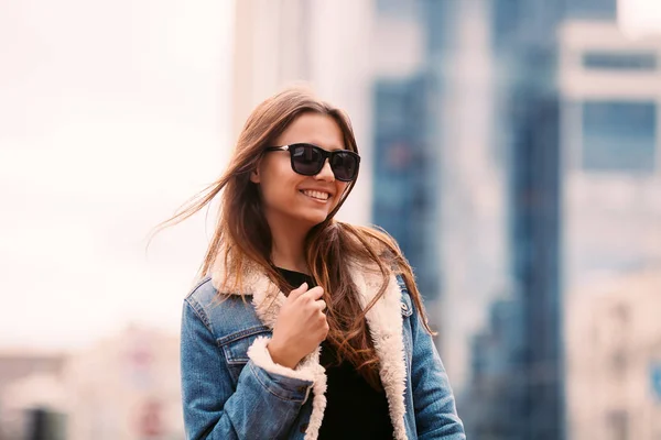 Kamera açık havada karşı bina gülümseyen uzun saçlı çekici kadın portre portre. Denim giyim, moda güneş gözlüğü takıyor — Stok fotoğraf