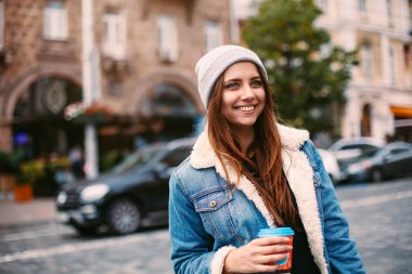 Kahve ile sokakta yürürken denim ceket uzun saçlı portre güzel kız gitmek. Kamera için heyecanlı görünüyor
