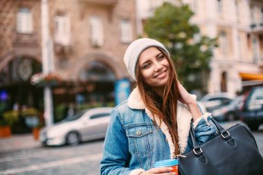 Şık hippi kız retro kot takım elbise ve örme şapka sokağın ortasında poz o dost gülümseyen, kahve tutar