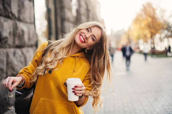 Stylish happy young woman wearing boyfrend jeans, white sneakers bright yellow sweetshot.She holds coffee to go. portrait of smiling girl in sunglasses and with bag — Stock Photo, Image