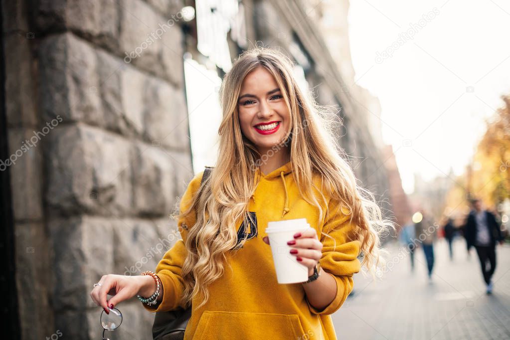 The concept of street fashion. young stylish girl student wearing boyfrend jeans, white sneakers bright yellow sweetshot.She holds coffee to go. portrait of smiling girl in sunglasses and with bag pos