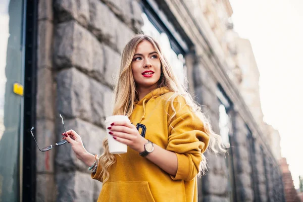 Pensive blond woman posing on modern streets, drinking coffee or cappuccino Street fashion concept, young woman wearing boyfrend jeans,glasses, white sneakers bright yellow sweetshot — Stock Photo, Image