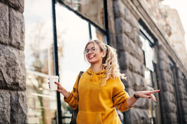 The concept of street fashion. young stylish girl student wearing boyfrend jeans, white sneakers bright yellow sweetshot.She holds coffee to go and dance . portrait of smiling girl in glasses — Stock Photo, Image