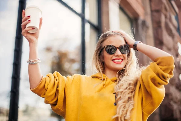 Eleganta glad ung kvinna som bär boyfrend jeans, vita sneakers ljusa gula sweetshot. Hon håller kaffe att gå. Porträtt av leende flicka i solglasögon — Stockfoto