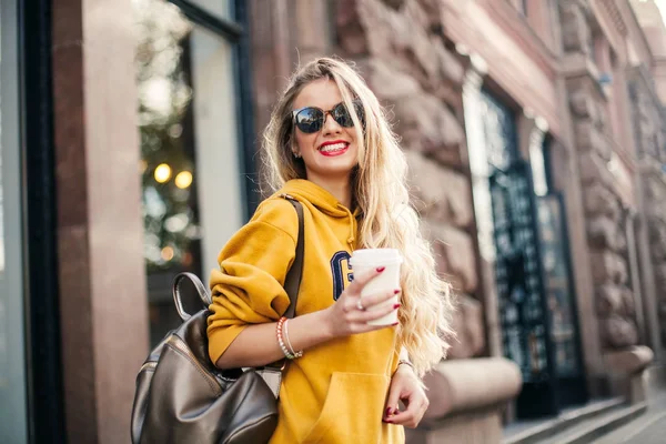 Unga vackra blonda dricka kaffe promenader runt city.mustard sweetshot., urban ryggsäck, klarröda läppar poserar mot fönstret i butiken — Stockfoto