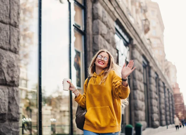 The concept of street fashion. young stylish girl student wearing boyfrend jeans, white sneakers bright yellow sweetshot.She holds coffee to go and dance . portrait of smiling girl in glasses — Stock Photo, Image