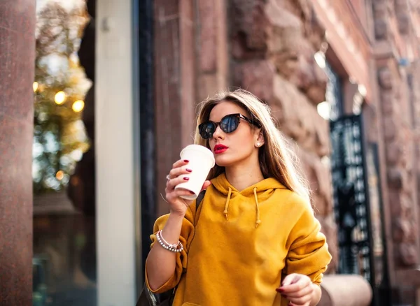 Stylish happy young woman wearing boyfrend jeans, white sneakers bright yellow sweatshirt.She holds coffee to go. portrait of smiling girl in sunglasses she greets friends — Stock Photo, Image