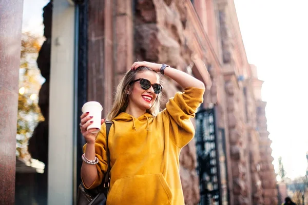 Eleganta glad ung kvinna som bär boyfrend jeans, vita sneakers ljusa gula sweetshot. Hon håller kaffe att gå. Porträtt av leende flicka i solglasögon och med väska — Stockfoto