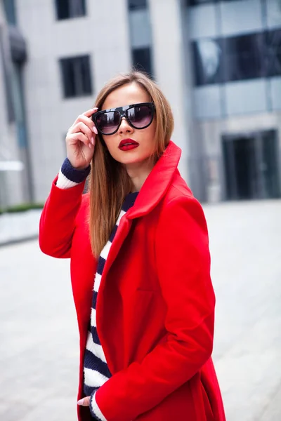 Joven chica con estilo, pasando por las ventanas.El uso de gafas de moda y un abrigo rojo. Resumen mirando hacia un lado. concepto de moda callejera — Foto de Stock