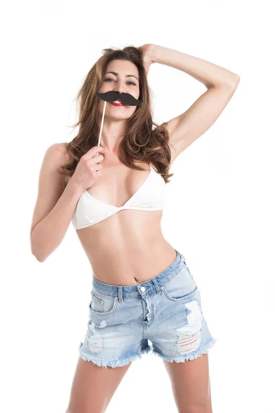 Attractive girl in jeans and bikini posing with paper moustache on wood stick, isolated on white background. Fun and joke concept