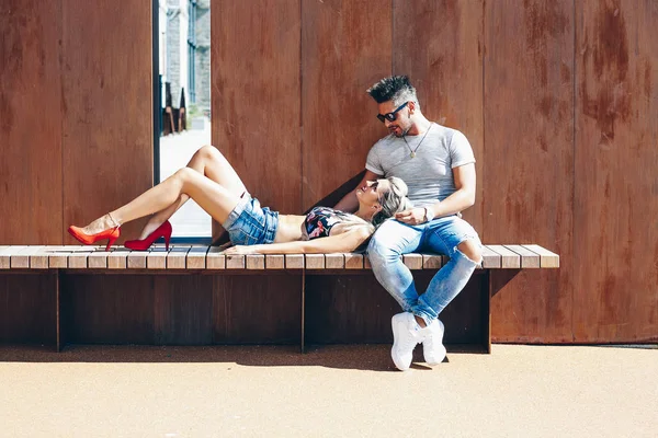 Portrait of handsome couple in casual clothing with sunglasses sitting on wood bench, joyful smiling expressions with heads together. Concept Loving Couple.