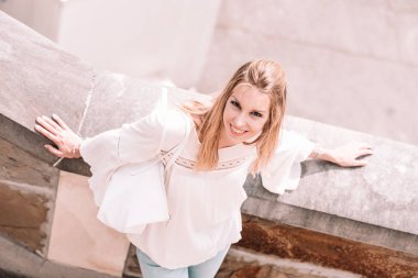 Beyaz bluzlu sarışın kadın çimento çitlere yaslanmış poz veriyor.