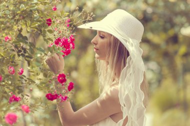 Beyaz şapkalı sarışın bir kadının portresi. Çiçekli bir köpek gülü çalısı.