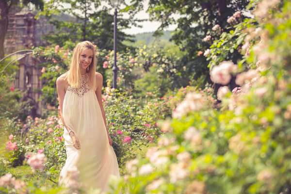 Smiling blonde woman walking in garden with blooming dog-rose bushes