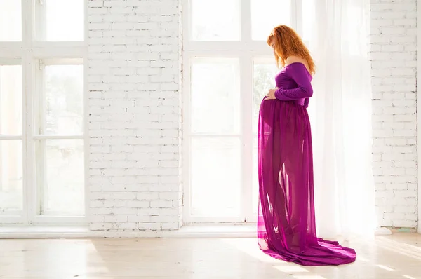 Беременная женщина в фиолетовом платье в белой комнате — стоковое фото