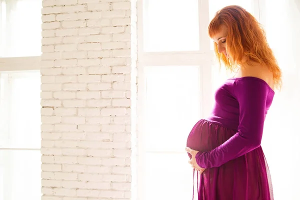 Mujer embarazada en vestido púrpura en habitación blanca —  Fotos de Stock