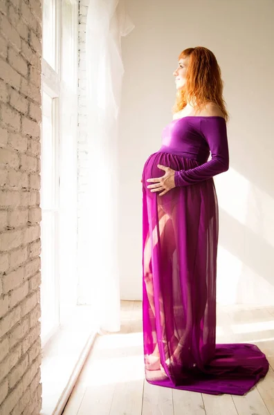 Mujer embarazada en vestido púrpura en habitación blanca —  Fotos de Stock