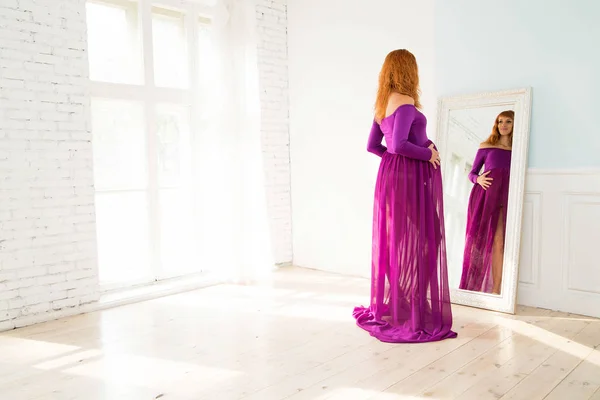 Pregnant woman in purple dress looks in mirror — Stock Photo, Image