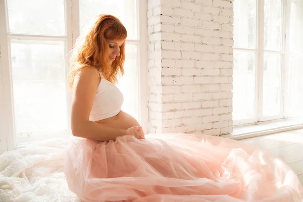 Zwangere vrouw in een taffeta rokje zit in de buurt van het venster — Stockfoto
