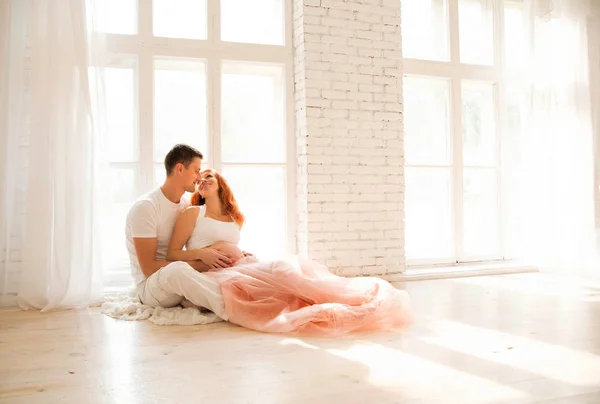 Mujer embarazada en una falda de tul y su marido están sentados en el suelo cerca de una gran ventana — Foto de Stock