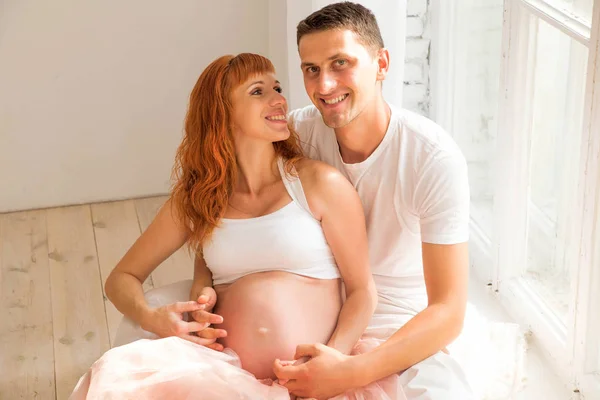 Kobieta w ciąży w tiulowym i jej mąż siedzi na podłodze w pobliżu duże okno — Zdjęcie stockowe