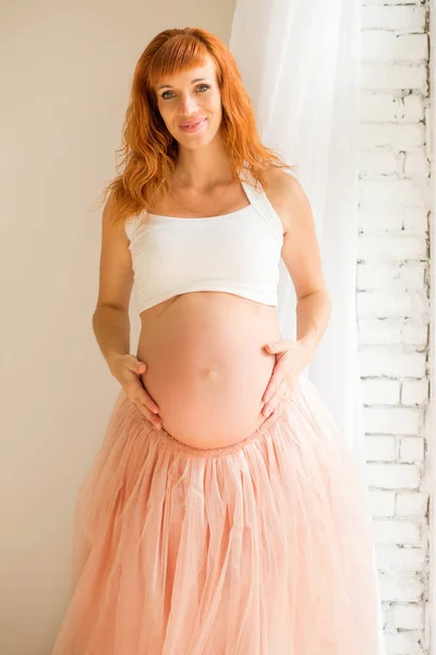 Een zwangere vrouw in een witte top en een Tule rok staat in de buurt van een groot raam — Stockfoto