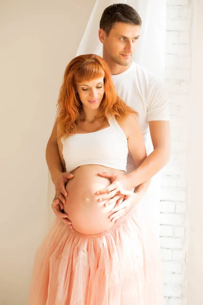 Una donna incinta con un top bianco e una gonna da smoking e suo marito stanno vicino a una grande finestra e godono del sole — Foto Stock