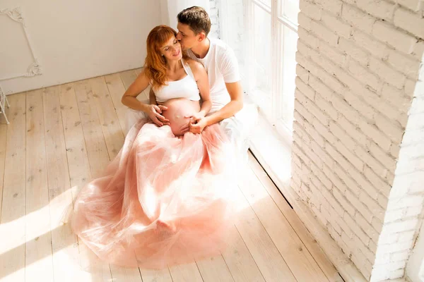 チュール スカートと彼女の夫の妊娠中の女性は、大きな窓の近くの床に座っています。 — ストック写真