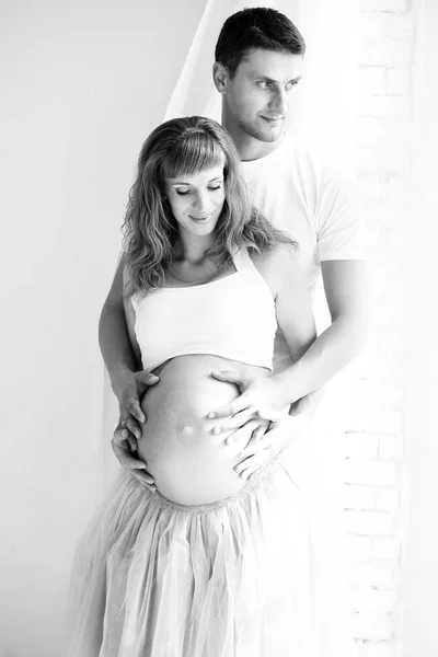 Kobiety w ciąży w biały top i Spódniczka smokingu i jej mąż stanąć w pobliżu duże okno i cieszyć się słońcem — Zdjęcie stockowe