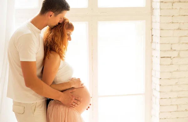 Une femme enceinte avec un haut blanc et une jupe en smoking et son mari se tiennent près d'une grande fenêtre et profitent du soleil — Photo