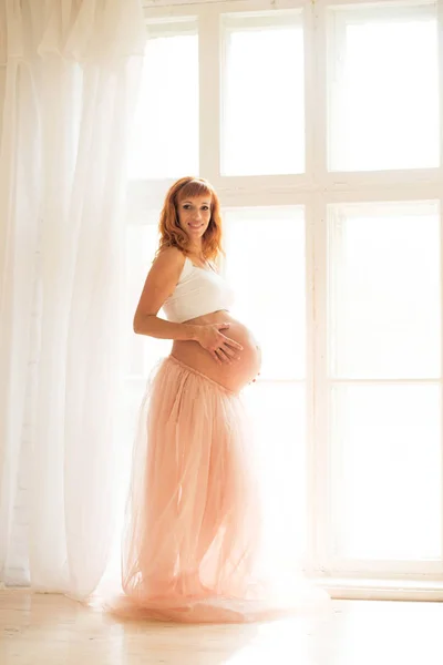 Een zwangere vrouw in een witte top en een Tule rok staat in de buurt van een groot raam — Stockfoto